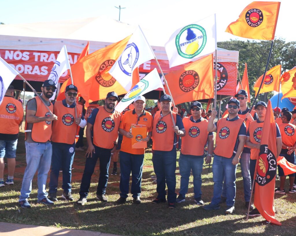 Trabalhadores do SECHSEG e Miguel Torres., presidente da Força Sindical, segurando bandeiras da força sindical e todos de colete laranja da força sindical na Marcha da Classe Trabalhadora.