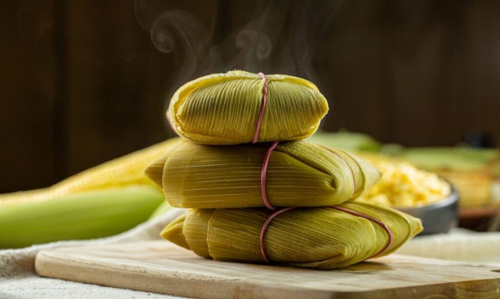 Imagem de mesa com tábua de madeira. Em cima dela têm três pamonhas saindo fumaça. no fundo está desfocado, mas há espigas de milho e palha.