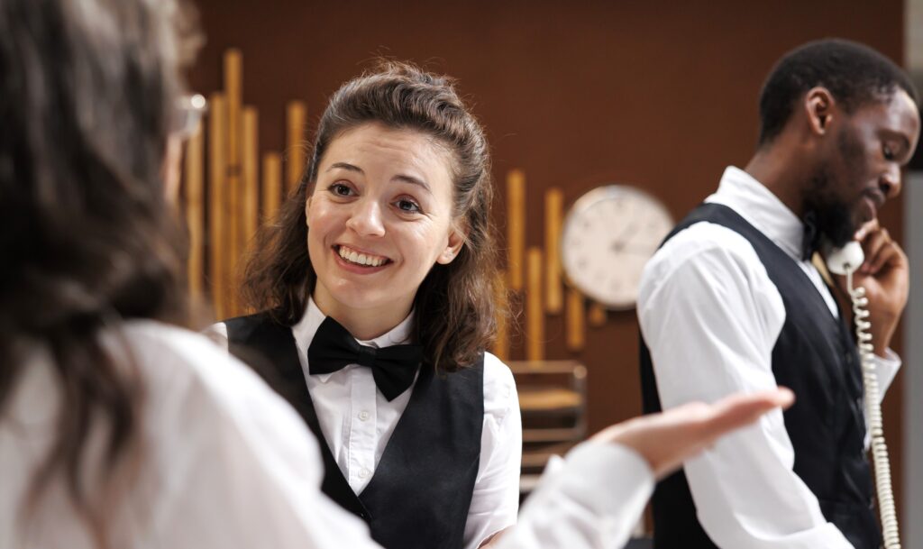 A imagem se passa em um hotel. Uma hóspede está falando com a funcionária que está sorrindo. Ao fundo um funcionário está fazendo uma ligação.