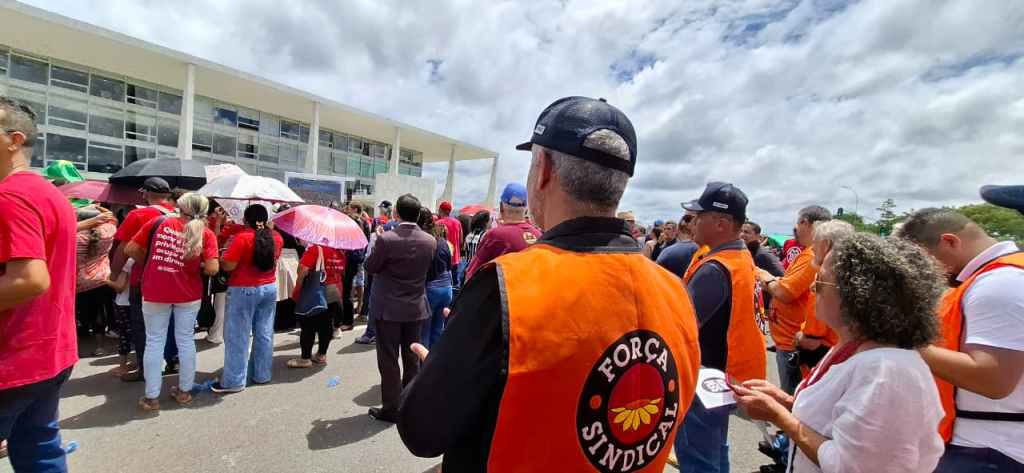 Pessoas assistindo telão de fora do Palácio do Planalto, em Brasília, transmitindo ao vivo a fala do Presidente Lula. Perto da câmera são os do SECHSEG usando o colete de força sindical.