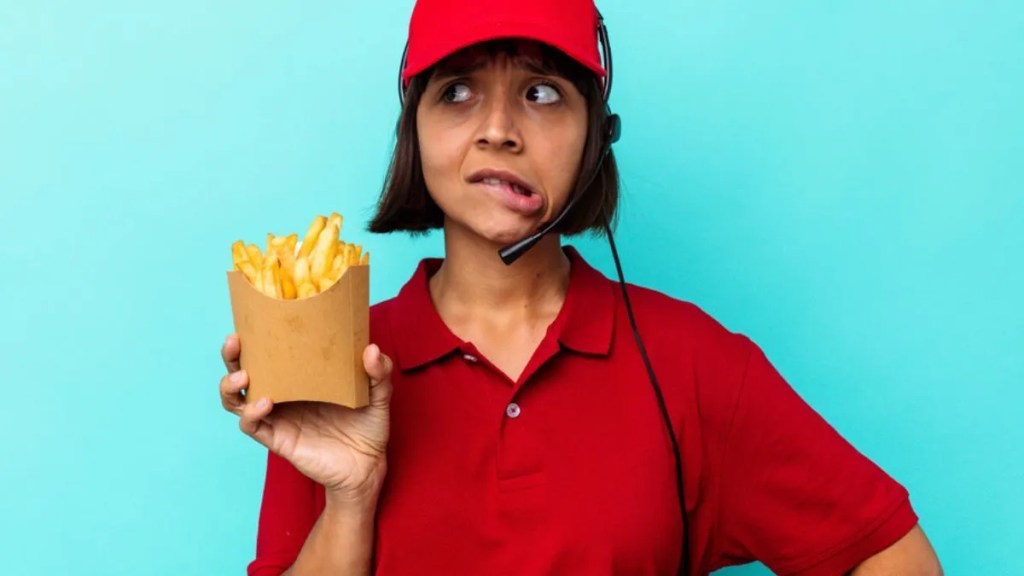 Atendente de fast-food com uniforme vermelho e microfone de drive-thru, está segurando uma embalagem com batatas fritas na mão e fazendo uma cara de preoucupada.