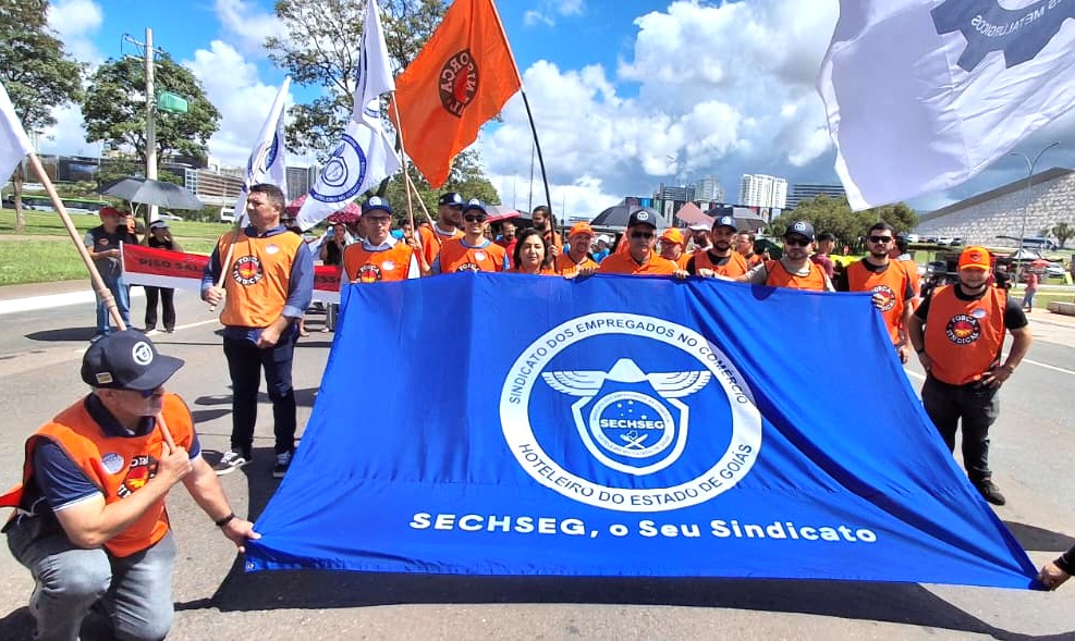 Equipe do Sechseg está usando coletes da Força Sindical e segurando uma grande bandeira azul do Sechseg.