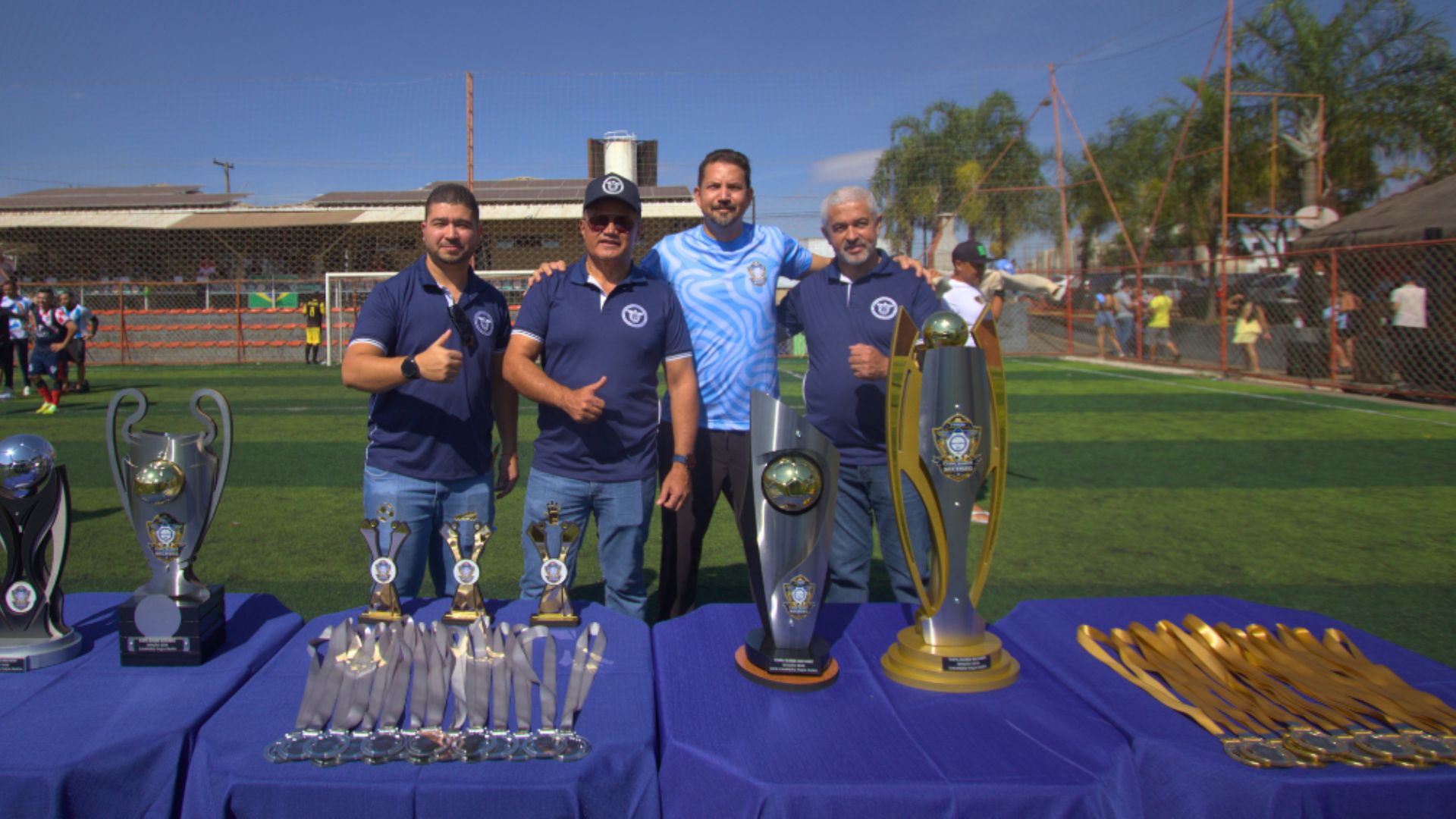 Presidente, vice-presidente e diretor financeiro do sindicato com arbitro em campo de futebol com mesa na frente com os troféus e medalhas.